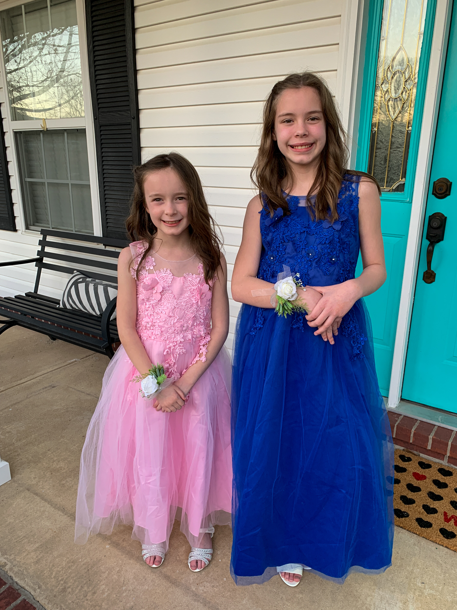 two girls stand in pink and blue party dresses with faux corsages on wrists