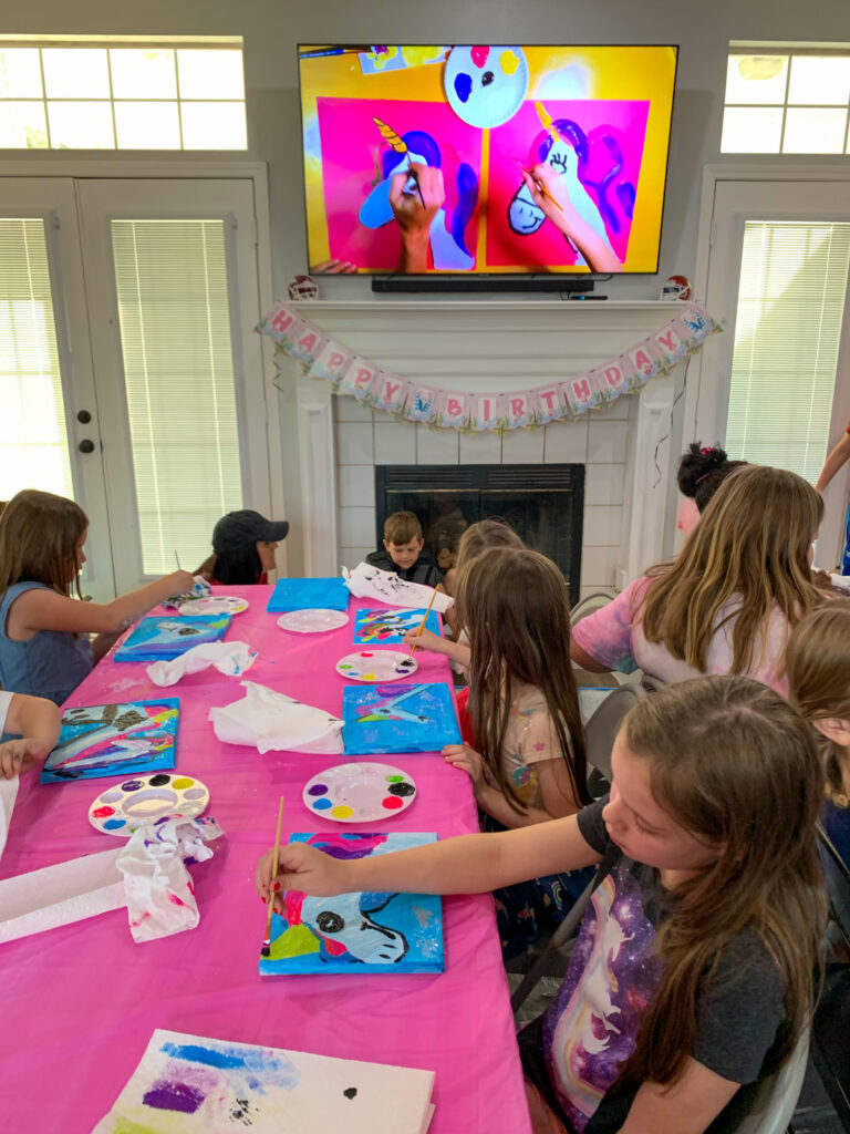 kids at a paint a unicorn party for a kids birthday with a TV screen tutorial above on mantle
