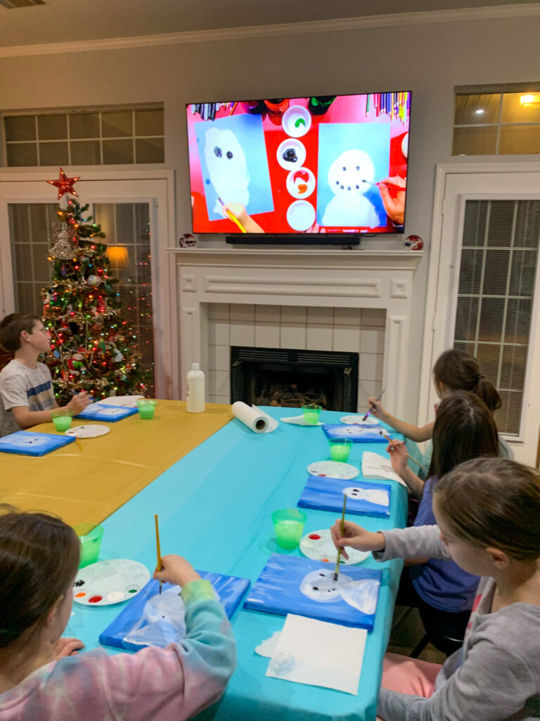 children watch a TV screen and paint a snowman on canvas at a snowman paint night at snowman themed party