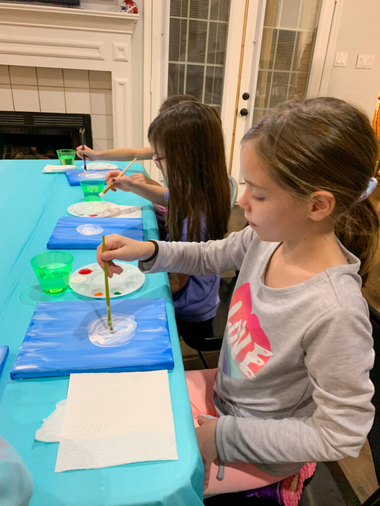 girls painting on a blue canvas at a snowman paint night